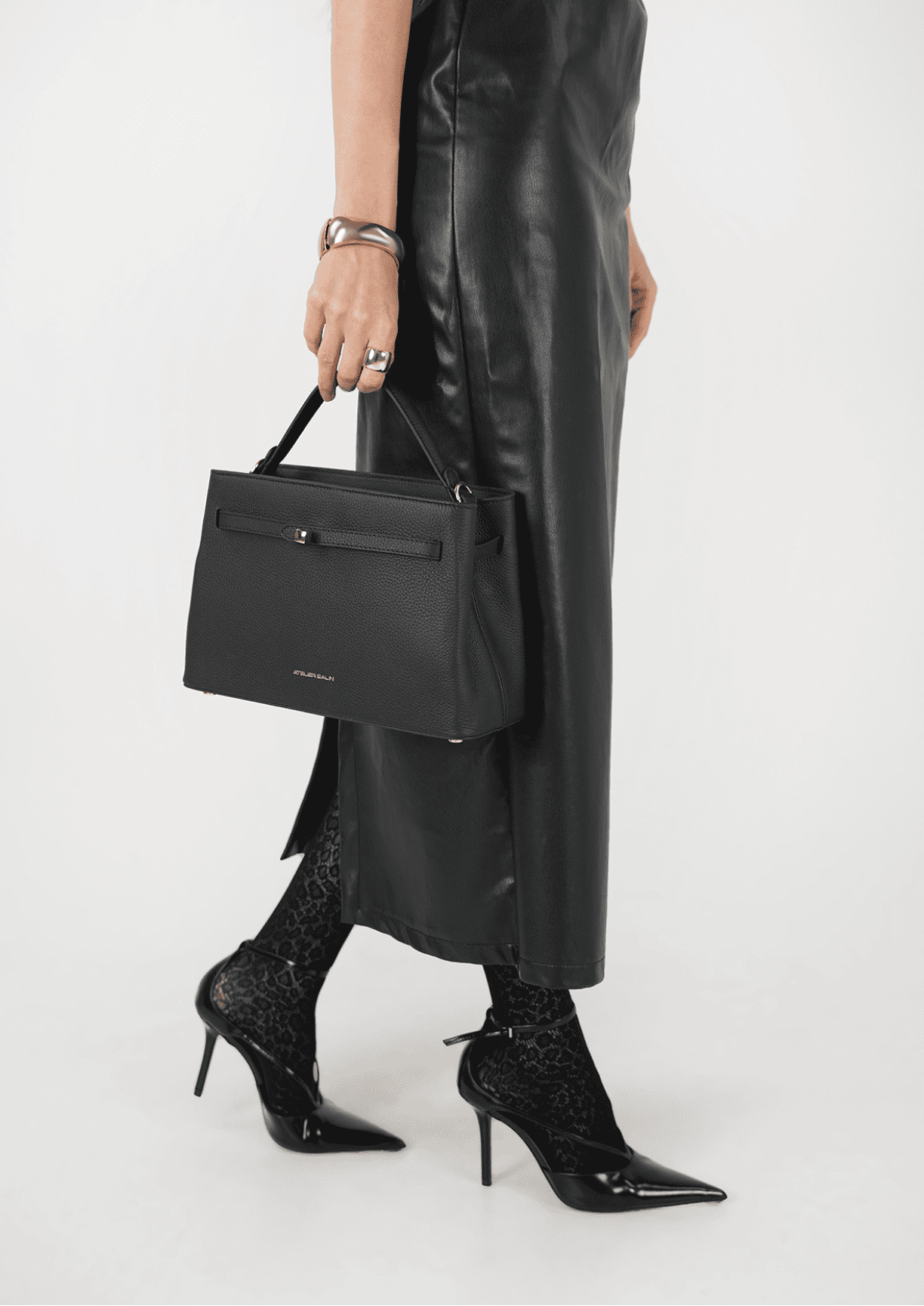 Person wearing a black leather outfit with a black leather atelier galin cube lock handbag on a white background