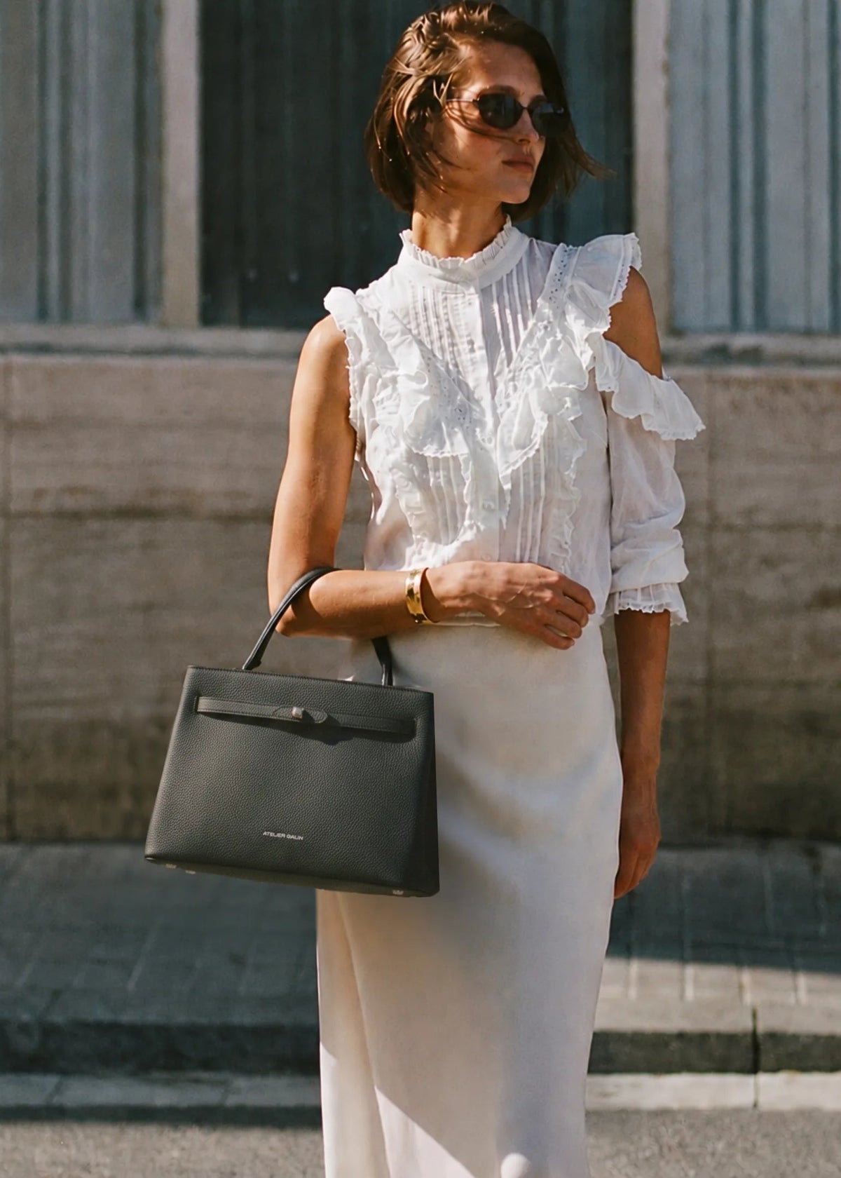 A woman wearing a white ruffled cold-shoulder blouse and a white midi skirt, carrying a black structured leather handbag, standing outdoors against a stone wall.