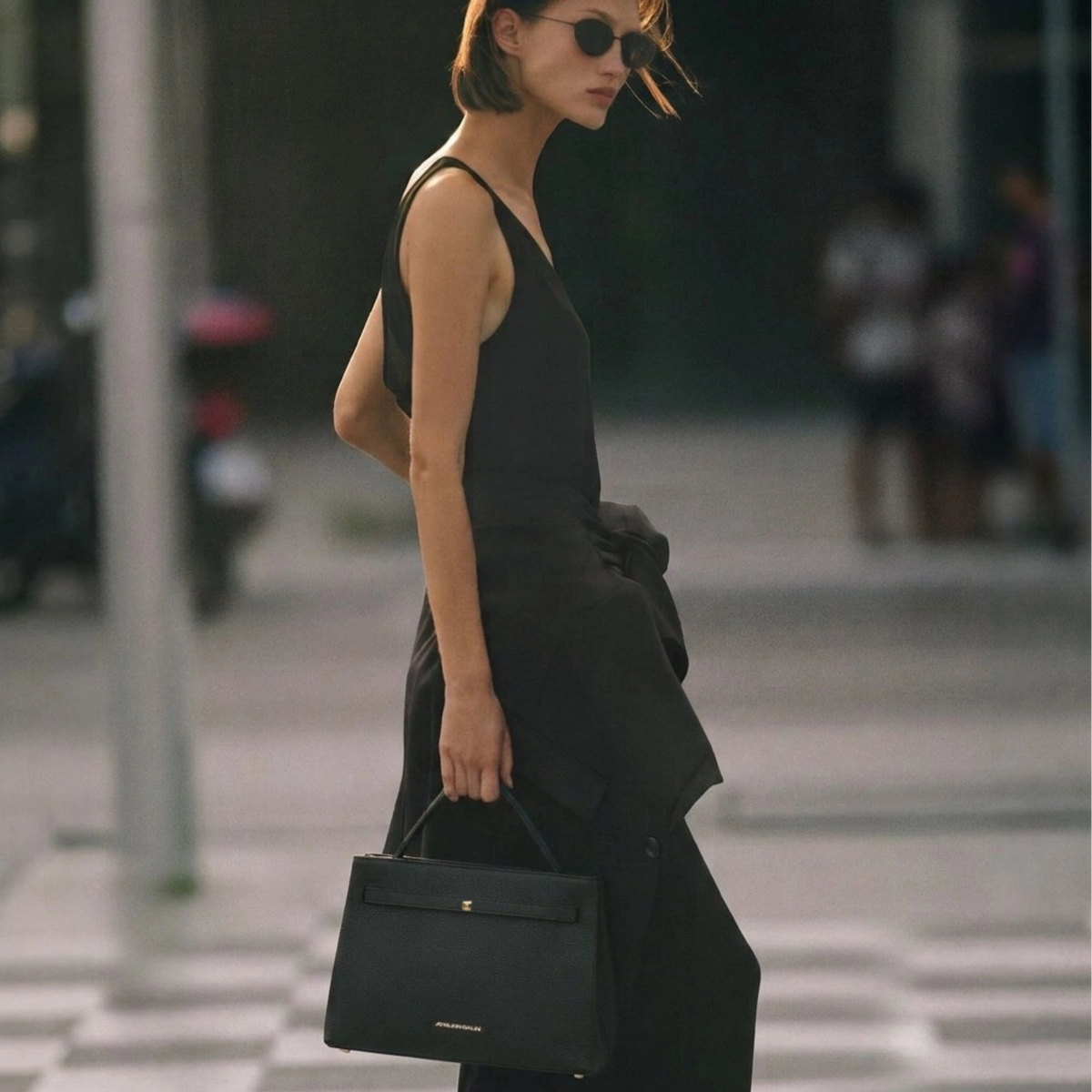 A woman in a black sleeveless top and wide-leg pants walks while carrying a structured black handbag with gold hardware.