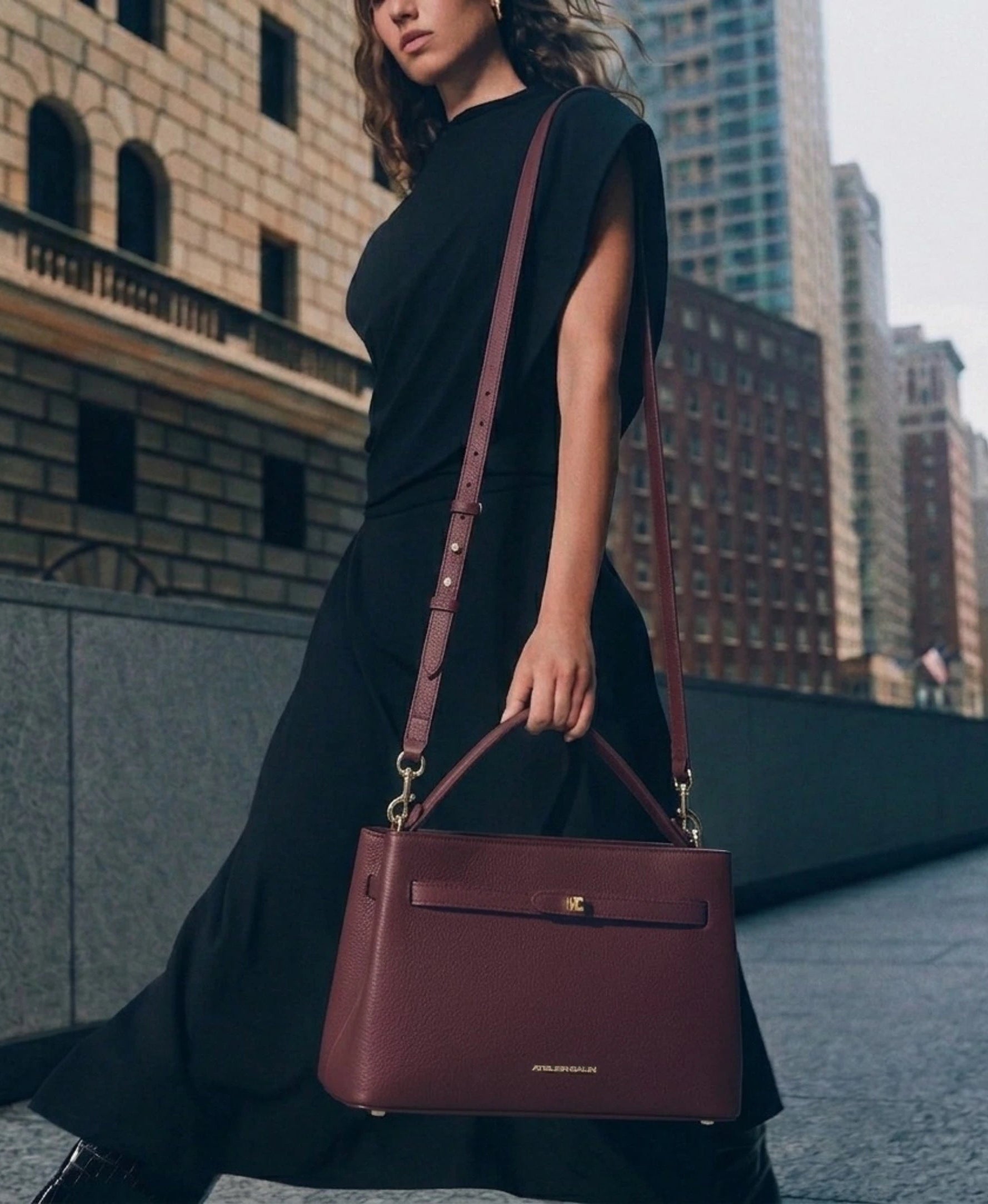 A woman in a black dress carries a structured burgundy leather handbag with a top handle and shoulder strap, walking along a city street with buildings in the background.
