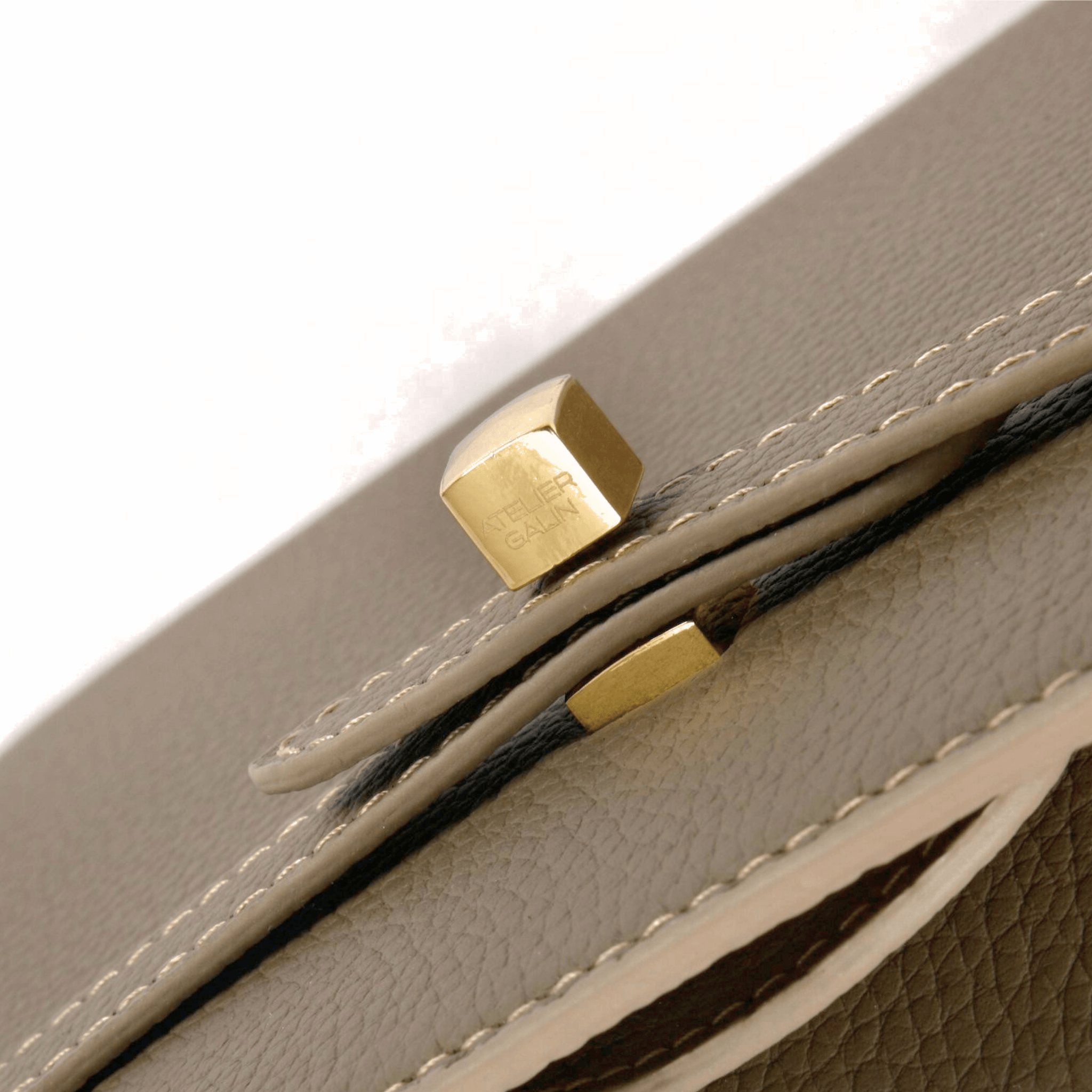 Close-up of a beige leather bag with gold atelier galin logo cube lock hardware on a white background