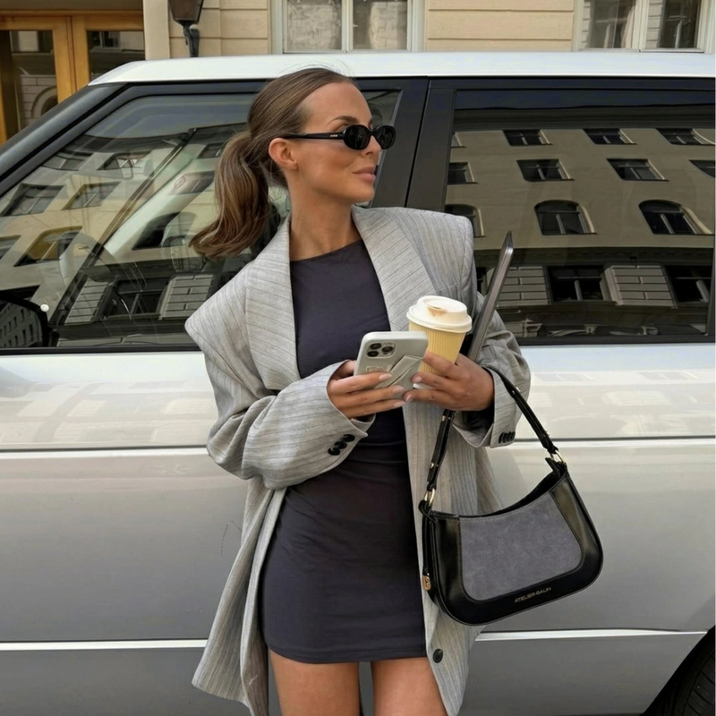 A woman wearing a light gray pinstripe blazer, dark gray mini dress, and black sunglasses stands in front of a silver car, holding a coffee cup and a black and grey suede shoulder bag.