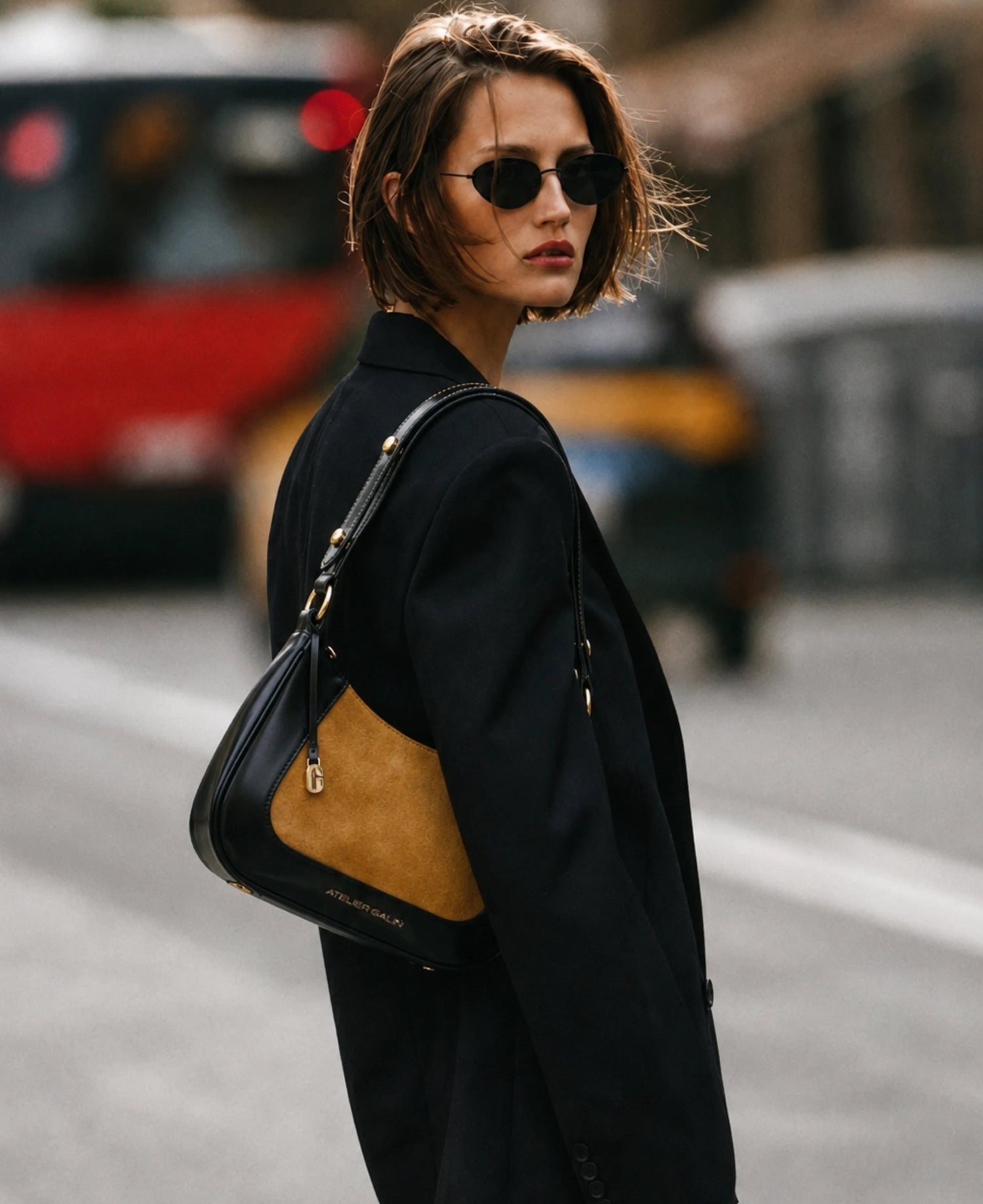 A woman wearing a black oversized blazer, black sunglasses, and carrying a black and tan leather shoulder bag with gold hardware, photographed on a blurred city street.