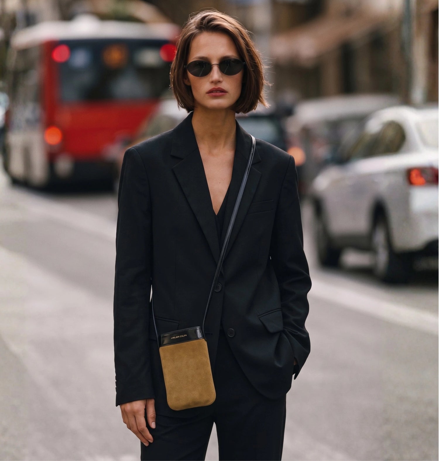 A woman wearing a black blazer and matching trousers stands on a city street, carrying a small suede crossbody phone bag pouch. She is wearing dark sunglasses.