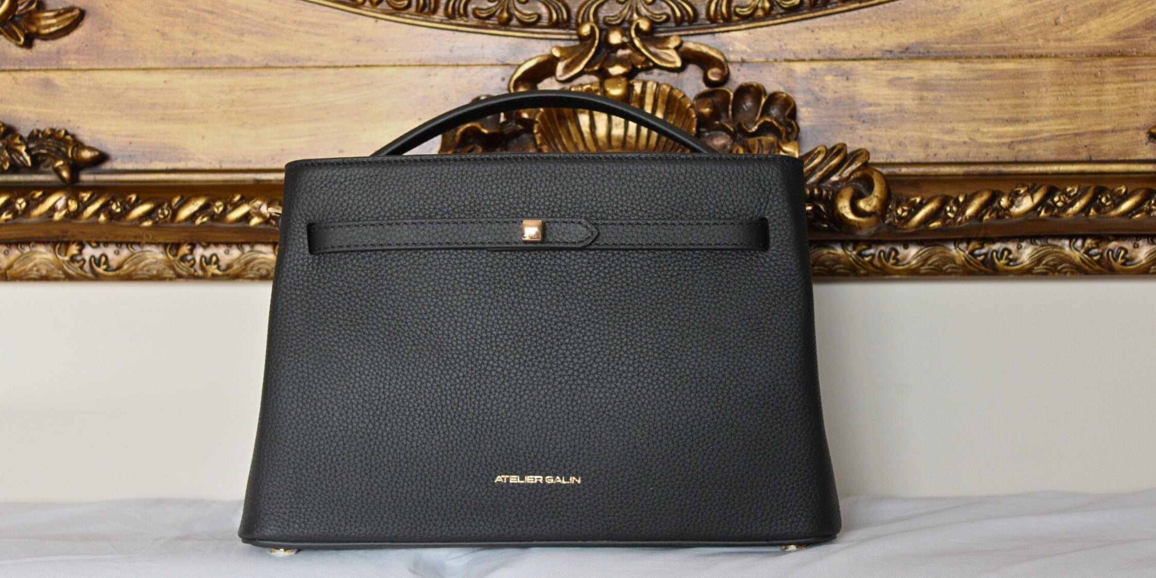 Black leather top handle handbag with cube lock and gold logo in front of an ornate mirror