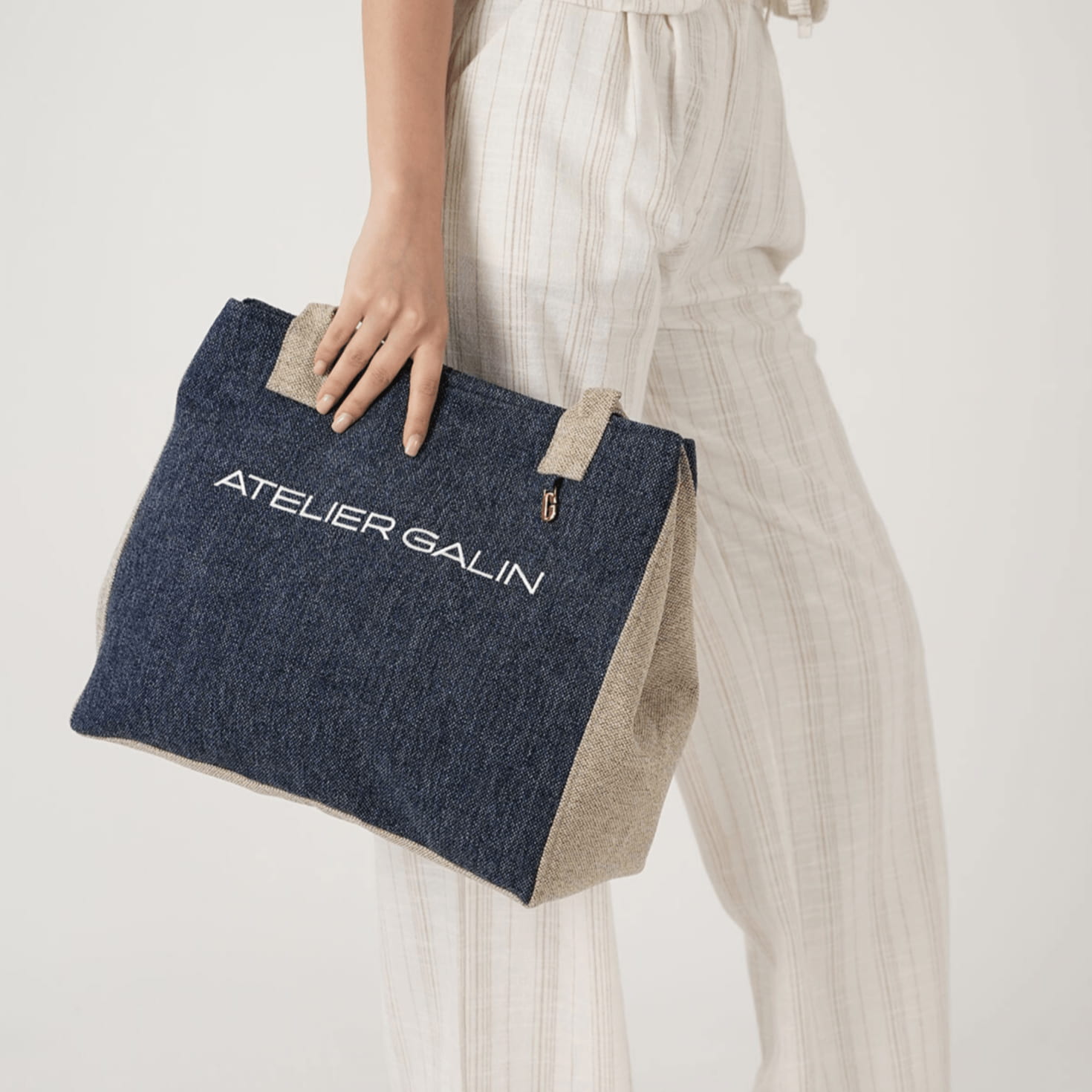 Person holding a blue and beige woven tote bag with 'Atelier Galin' branding on a light background